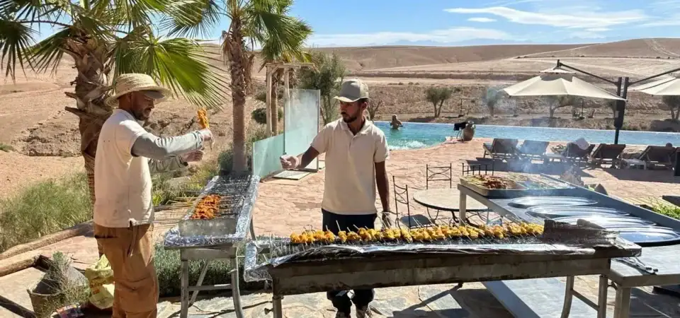 Grilled skewers being prepared at Marrakech Stay Vip, set amidst a desert landscape with palm trees and a luxurious pool area, offering an authentic Moroccan gastronomic experience.