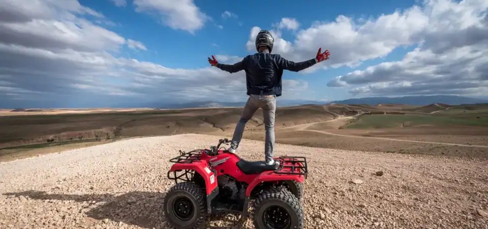 Vibrant image of a man confidently standing on a red ATV in a vast desert landscape with rolling hills and a partly cloudy sky, emphasizing adventure and luxury stays in Marrakech.
