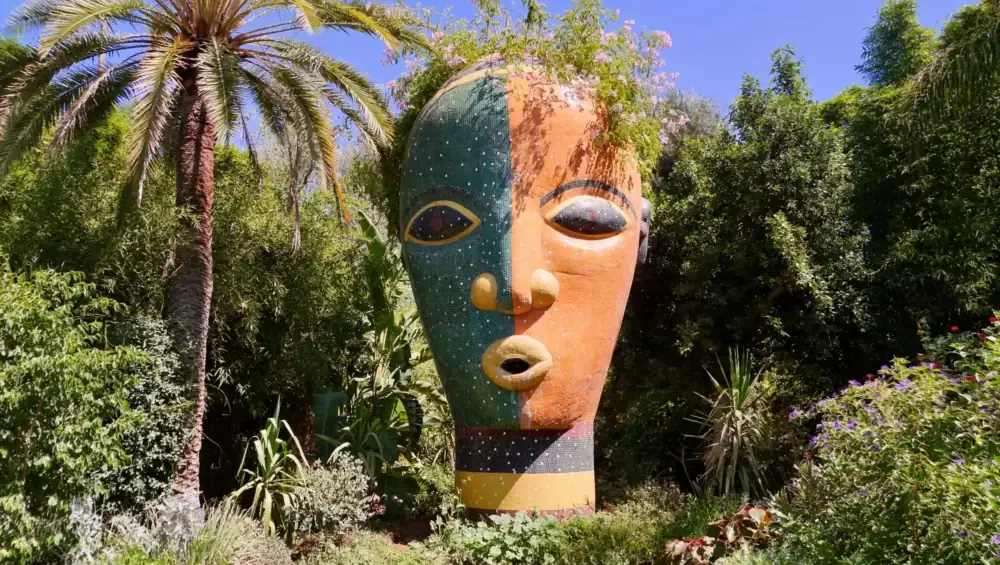 Vivid Moroccan garden with a large colorful sculpture of a face surrounded by lush greenery and palm trees, showcasing Marrakech Stay Vip's vibrant outdoor attractions.