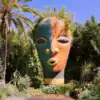 Vivid Moroccan garden with a large colorful sculpture of a face surrounded by lush greenery and palm trees, showcasing Marrakech Stay Vip's vibrant outdoor attractions.