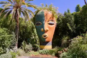 Vivid Moroccan garden with a large colorful sculpture of a face surrounded by lush greenery and palm trees, showcasing Marrakech Stay Vip's vibrant outdoor attractions.